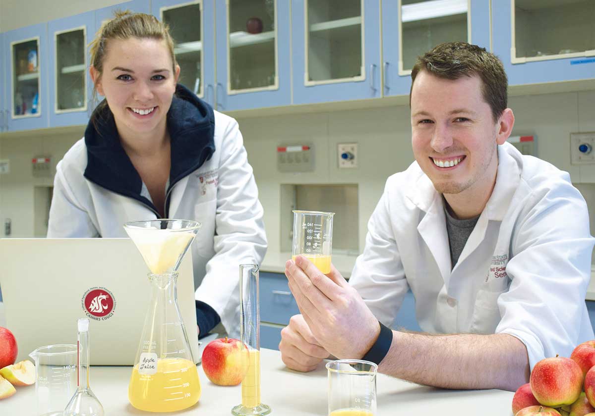 Two students in a lab, one with a laptop computer on the counter and the other with a beaker filled with yellow liquid.