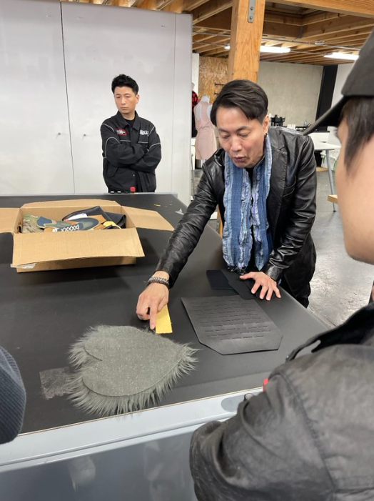 A Tuka Tech representative points towards a heart-shaped cutout of fabric with furry texture while talking to the students about how it's cut. Students stand in the background observing.