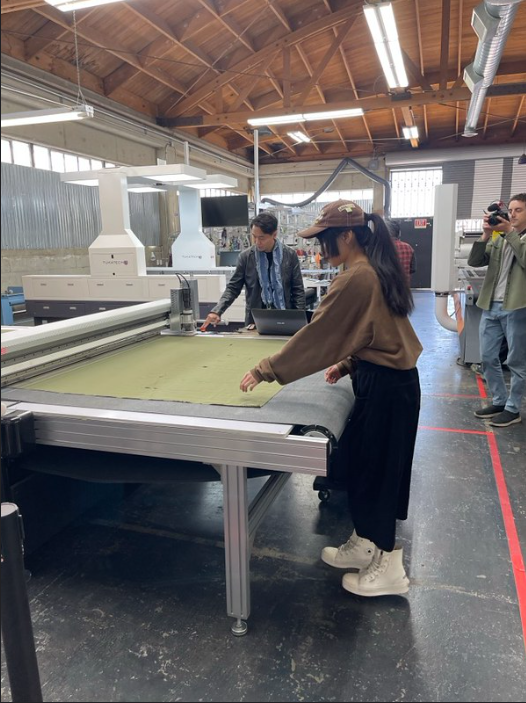 A student is seen laying her hands on an enormous conveyer belt that is feeding fabric through a laser cutter, with the head representative of Tuka Tech pressing buttons on the machine in the background.