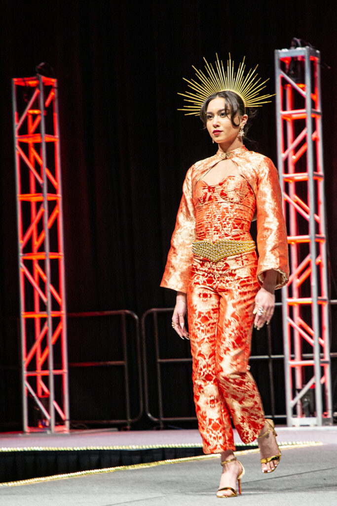A celestial look in vibrant orange brocade, the model stands tall with the confidence of the sun, a headpiece representing rays of light radiating from her head. A gold belt and gold shoes accessorize the look.