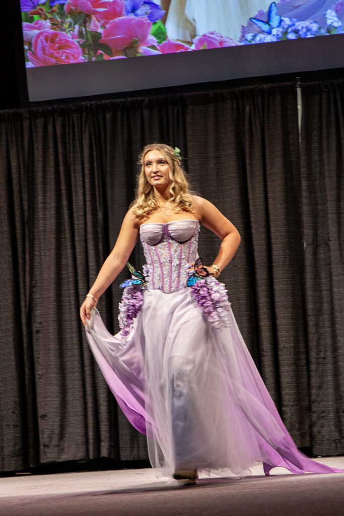 A floral lavender look, this dress features ruffled flowers and butterflies on the hips of a lace corset bustier. A long ombre train of floaty fabric trails behind her.