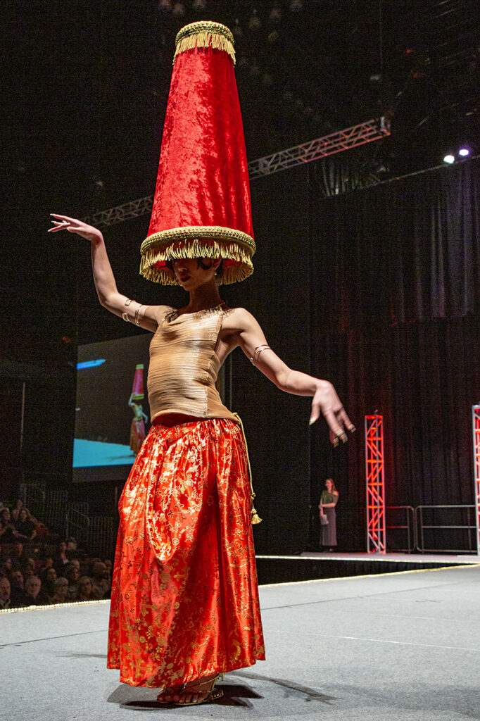 A model poses dramatically with fanciful arms adorned with bangles, wearing a highly-saturated red skirt with gold shining embroidery, a textured top, and a gigantic tall fringed lamp shade on top of her head.