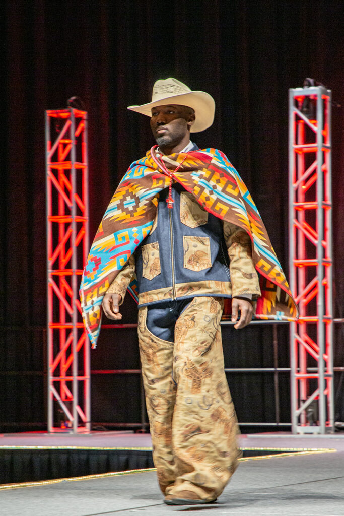 A fanciful cowboy look with intricate mixed patterns and denim is topped off with a cowboy hat, as the model is framed by bright lights on the runway.