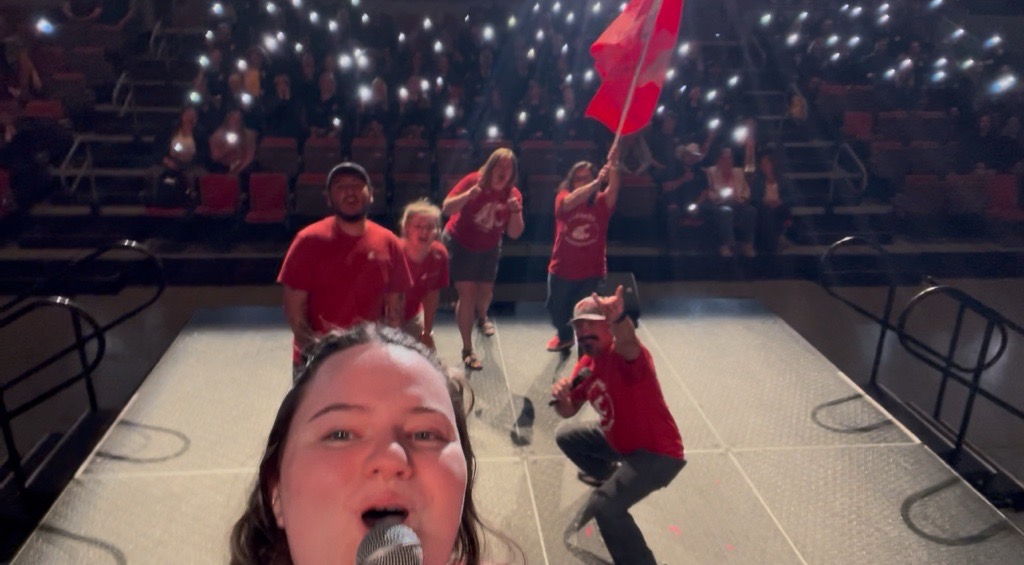 Colette Casavant, Miguel Inzunza, Emily Libey, and Jose Perez-Olmos wave a huge coug flag in front of a sea of students who are waving their phone lights and shouting Go Cougs.