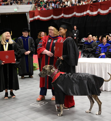 Wendy Powers stands next to a student who has her large dog with her. The dog is also dressed in a graduation gown.