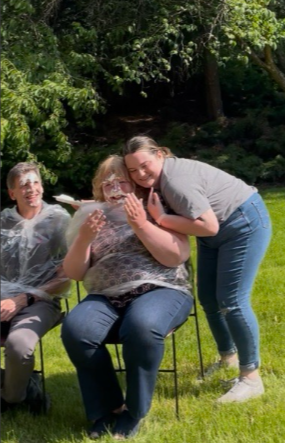 Emily hugging Dean Wendy Powers kindly while Wendy tries to wipe enough whipped cream off her eyes to see again. Scot smiles in the background.