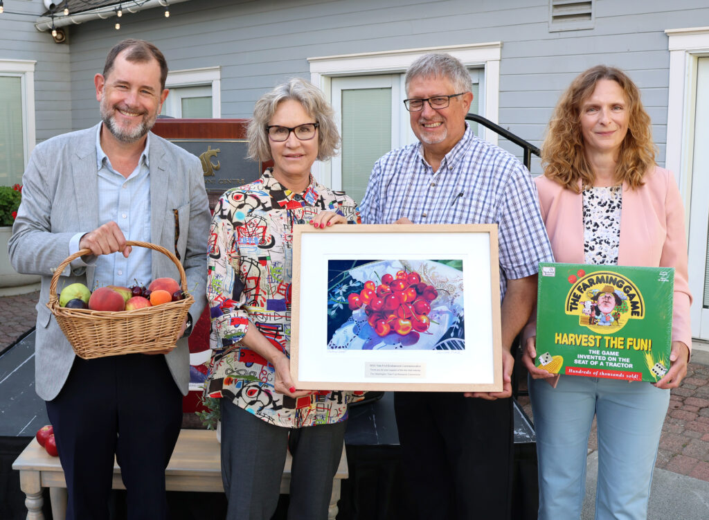 The President of WSU, Elizabeth Cantwell, The Provost, Chris Riley-Tillman, and harvest growers including Director of the Washington Tree Fruit Research Commission (WTFRC), Ines Hanrahan, pose with a basket of fruit and some commemorative art in front of the Alumni Center.