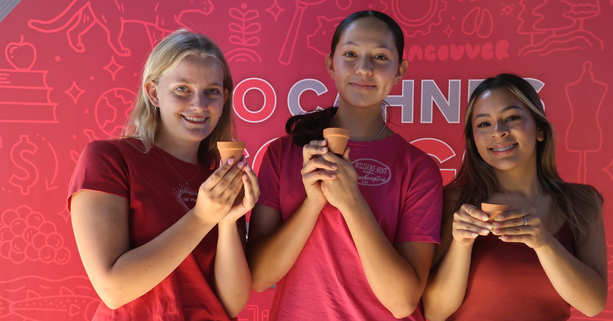 Three CAHNRS students in matching CAHNRS red t-shirts hold their tiny potted plants, smiling, in front of the CAHNRS background at a student Week of Welcome event.