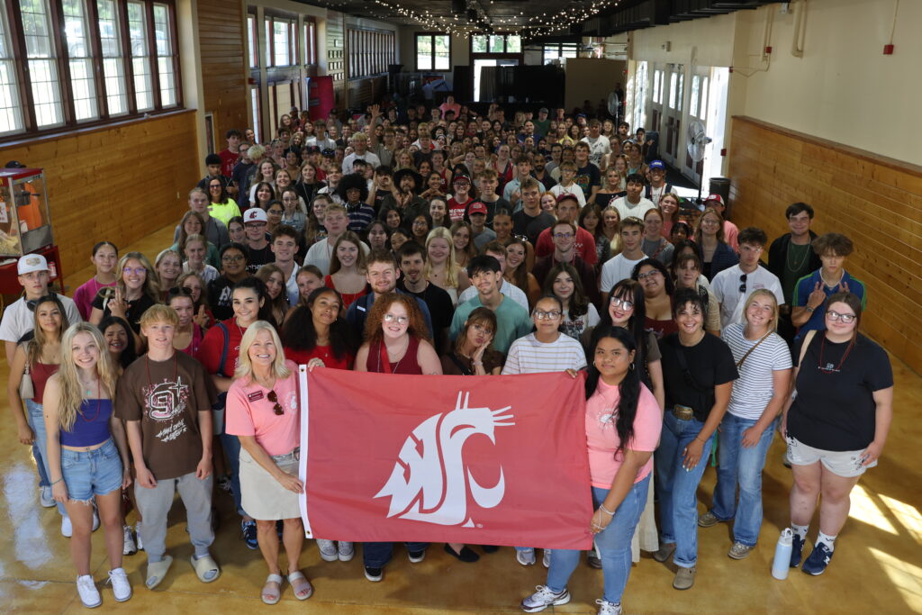 Hundreds of students stand behind Nancy Deringer and an Ambassador, holding the WSU flag at WOW Ack.