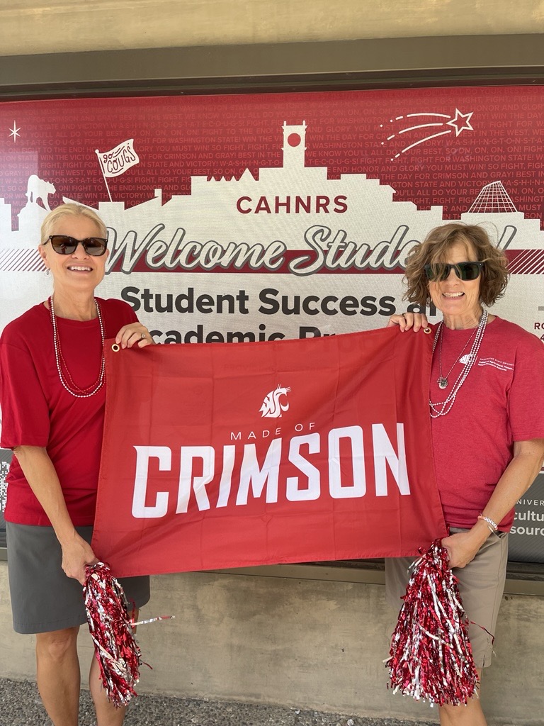 Nancy Deringer and Judy Hopkins hold the "Made of Crimson" flag and pom-poms in front of the CAHNRS Student Success and Academic Programs window.