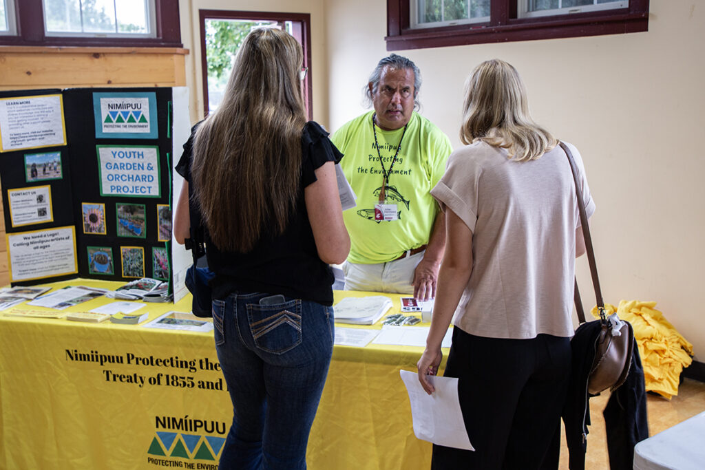 the Nimiipuu Youth Garden and Orchard Project representative explains his program to attendees at his booth with a colorful posterboard, reaching for a brochure