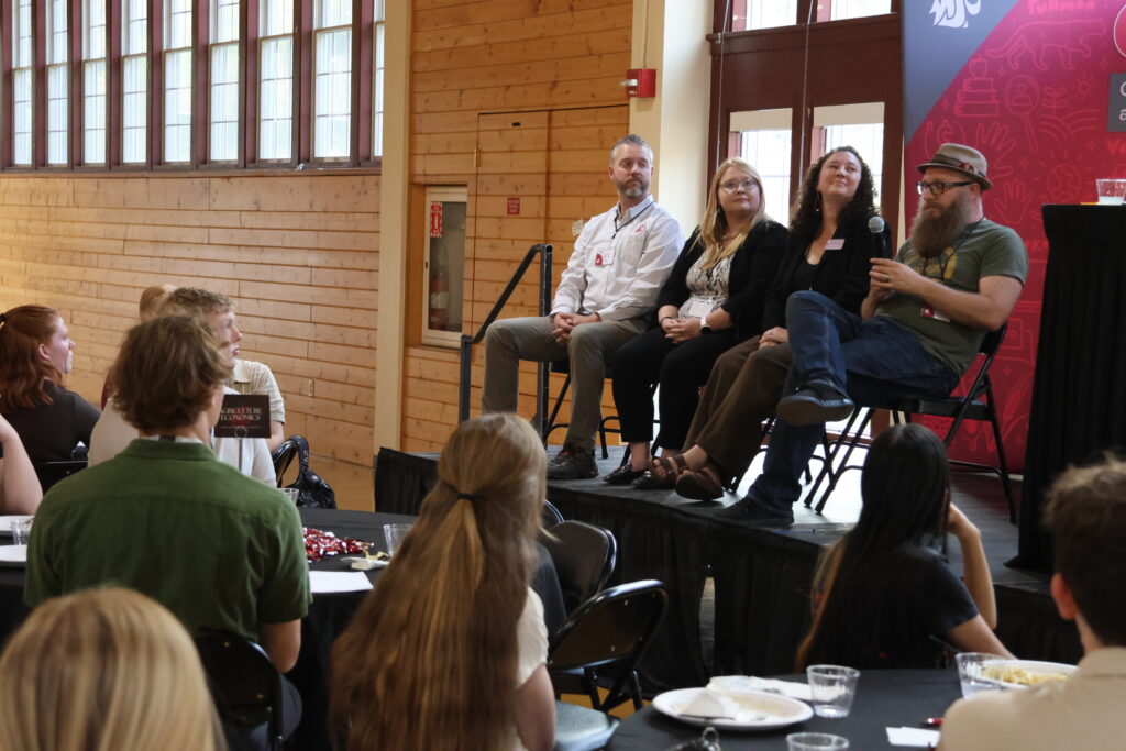 Mentors sit atop a stage at Orientation Dinner for the Top Scholars, speaking into a microphone about their advice for attendees, who sit listening attentively