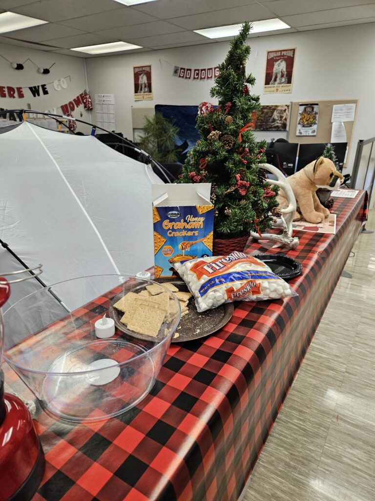 S'mores ingredients and an evergreen tree sit next to some antlers on a booth in the office of research.