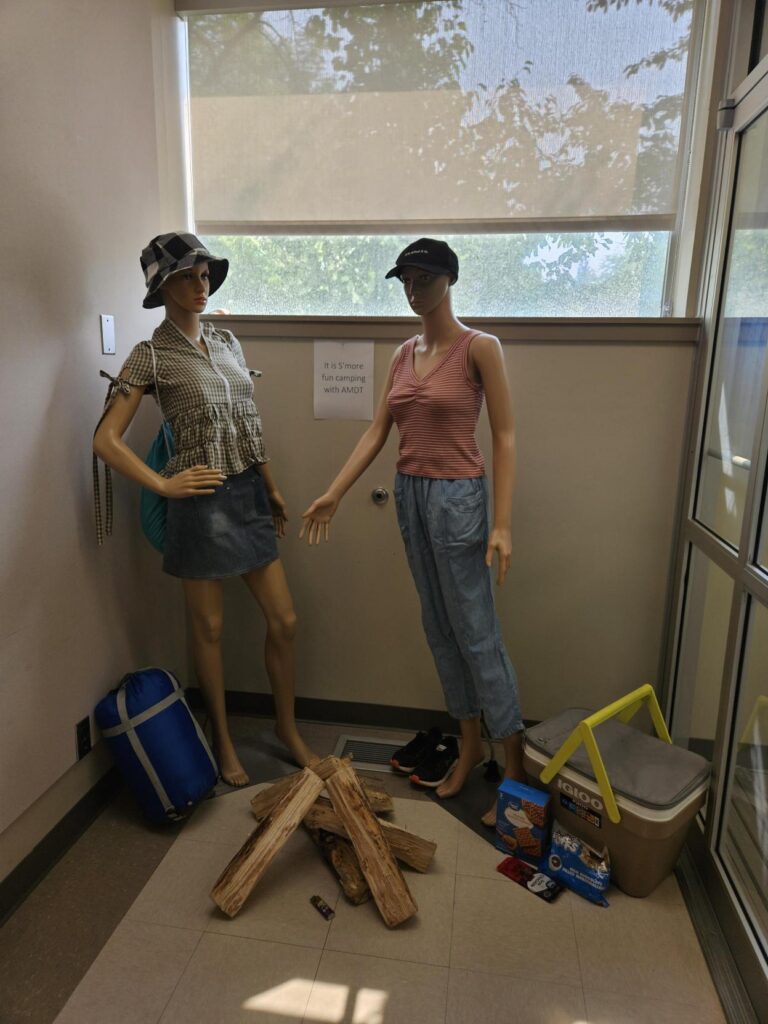 Two AMDT mannequins in camping outfit stand in front of s'mores ingredients and coolers, with a sleeping bag and a fire next to them