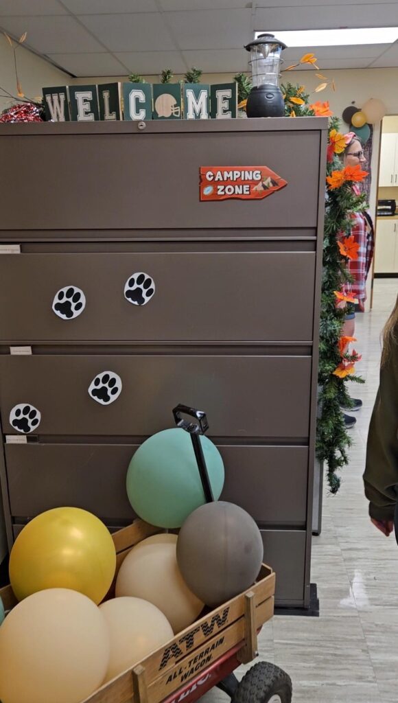 Baloons in a wagon lead to cougar paw marks on a filing cabinet, with the sign "Camping Zone"