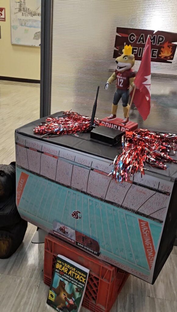 an elaborately drawn football field is shown on a tv made of cardboard with last year's first place trophy of Butch on top. The book "how to survive a bear attack" is propped up under there