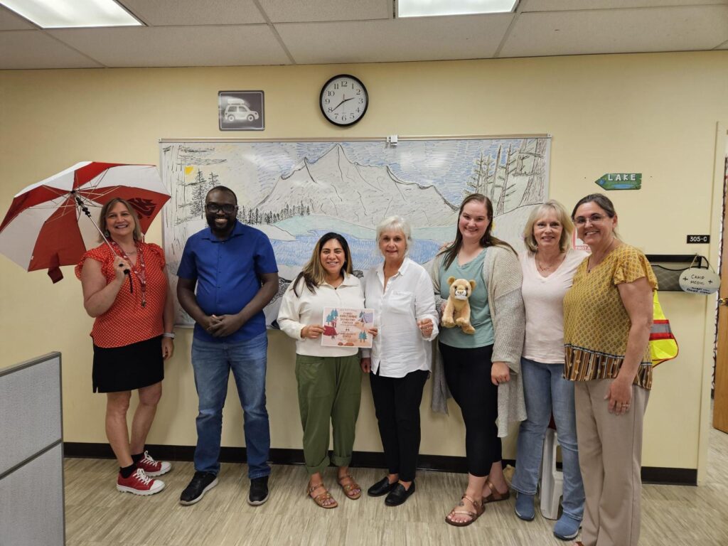 The Business Center team accepts their cougar plushie award and certificate for winning the homecoming decorating contest. The background includes an elaborate drawing of a mountain scene done in whiteboard marker, and one attendee holds a wsu umbrella
