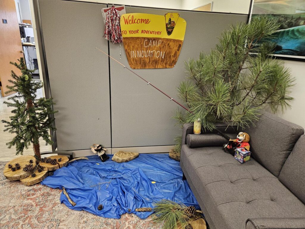 Welcome to Camp Innovation is written by trees and a woods scene in the office of research. A mini cougar plushie is fishing for a Vandal plushie in a pool made of fabric.