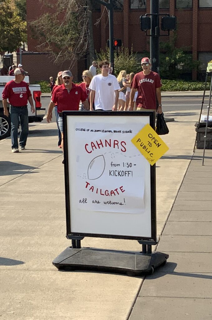 A sign says "College of Agricultural Human and Natural Resource Sciences - Open to Public: CAHNRS Tailgate - from 1:30 to kickoff: All are welcome" with a drawing of a football.
