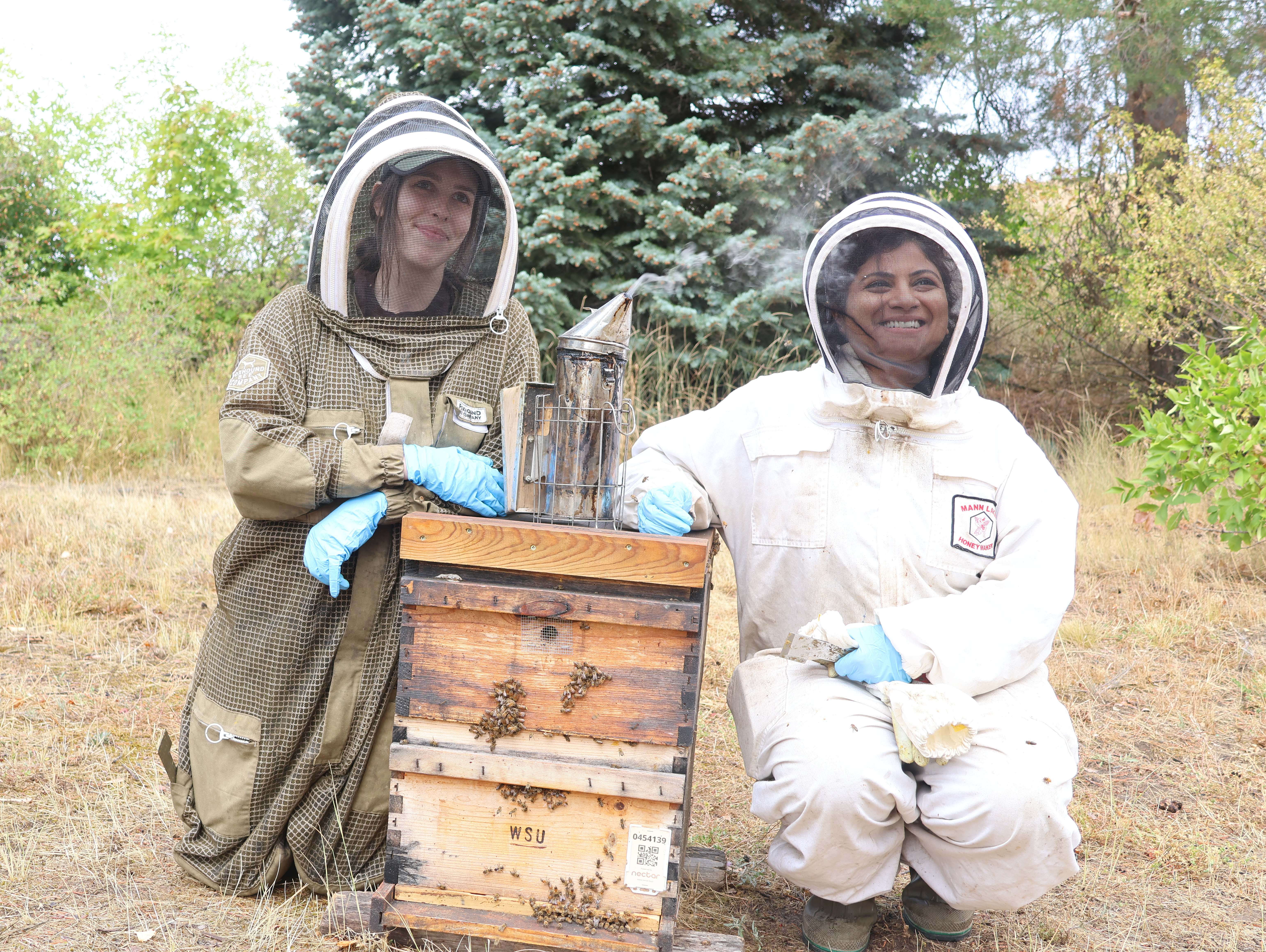 Two Entomology students in full protective gear kneel beside bee pollinator equipment during a class visit to the aviary
