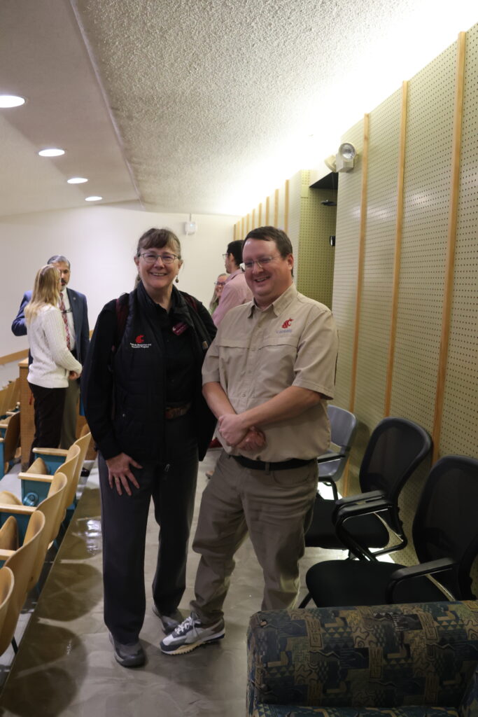 Two attendees of the All CAHNRS Meeting pose in the back of the classroom after it's finished. The dean is answering a question behind them.