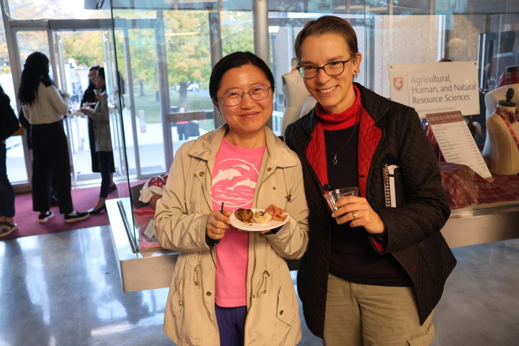 Two attendees of the CAHNRS Meeting Reception hold their drinks and snacks in front of the display case of AMDT tartan merchandise in Johnson Hall.