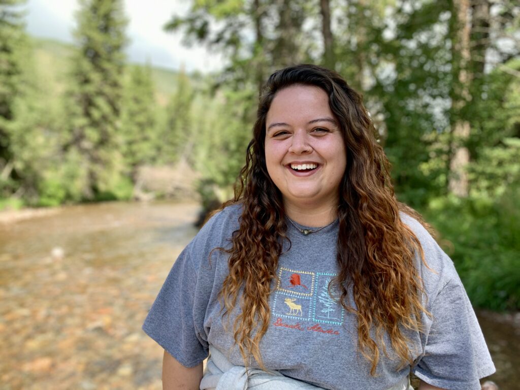 Aspen Dobbins smiles warmly on an adventure in the outdoors with the trees behind her. She wears a forest-inspired t-shirt.