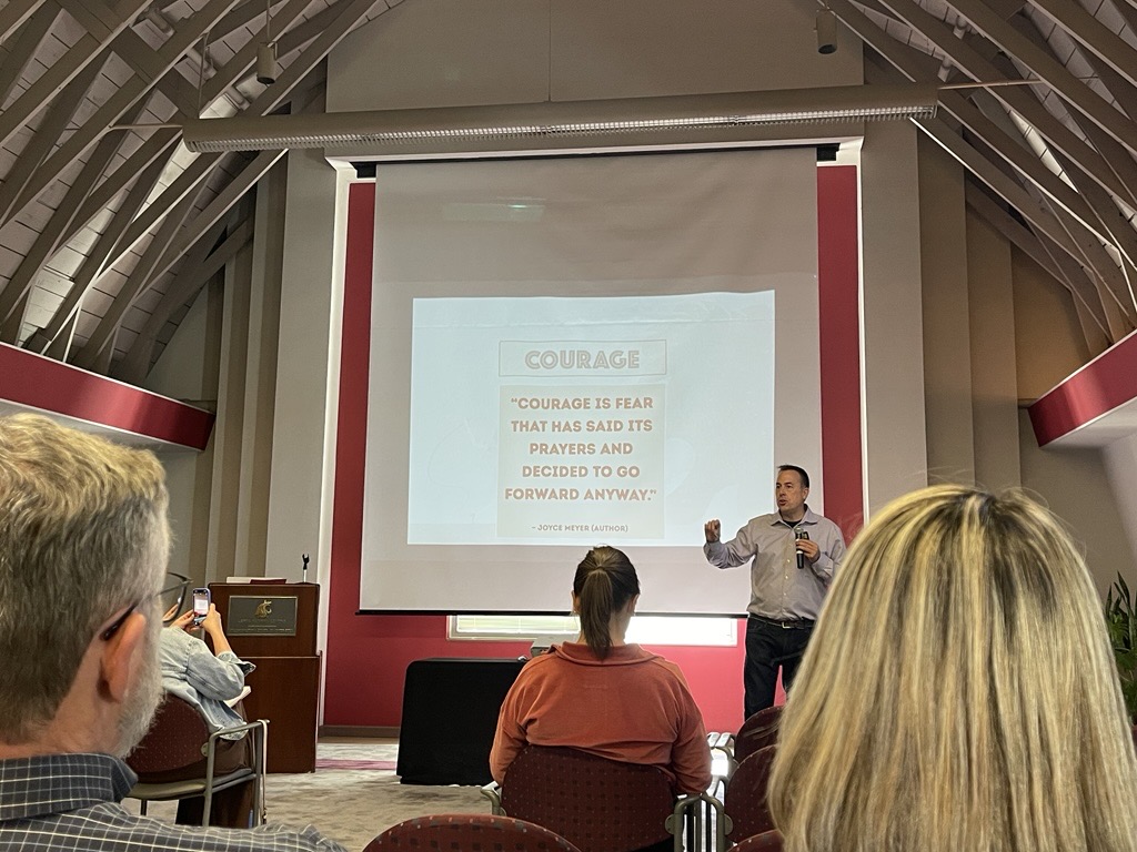 Corey Ciochetti animatedly waves his arms, making a point about courage and fear. The slide behind him reads " Courage is Fear that has said its prayers and decided to go forward anyway"