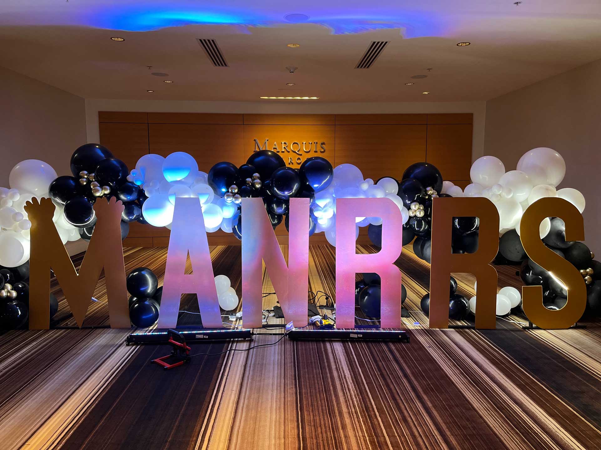 an arch of balloons and the giant cutout letters that spell out MANRRS are illuminated in iridescent light