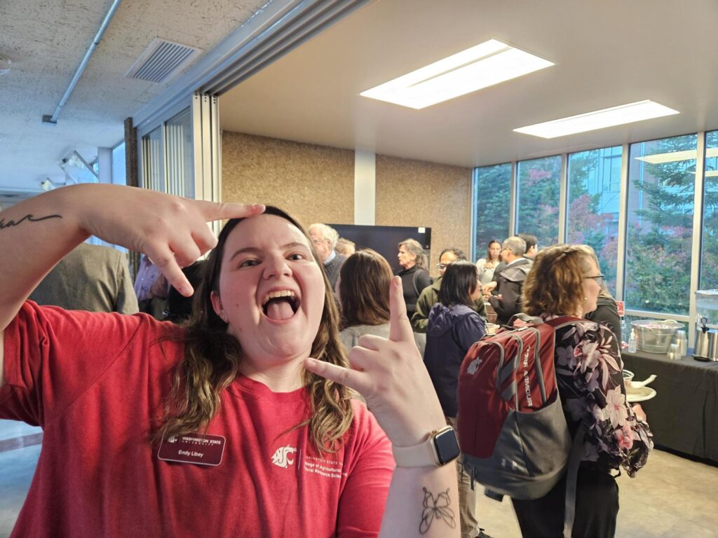 Emily Libey making a silly pose, wearing a red CAHNRS tshirt at the All-CAHNRS Reception. Behind her are tables of food and other attendees.