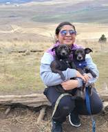 Nancy Hernandez holds two dogs and sports fabulous sunglasses, kneeling in nature.