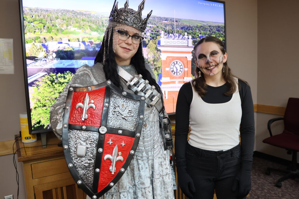 Kat Tsatsomeros wears a silver chainmail dress, chainmail hood, and crown, holding a shield. Jorja Heinkel wears a black and white outfit with skull makeup.