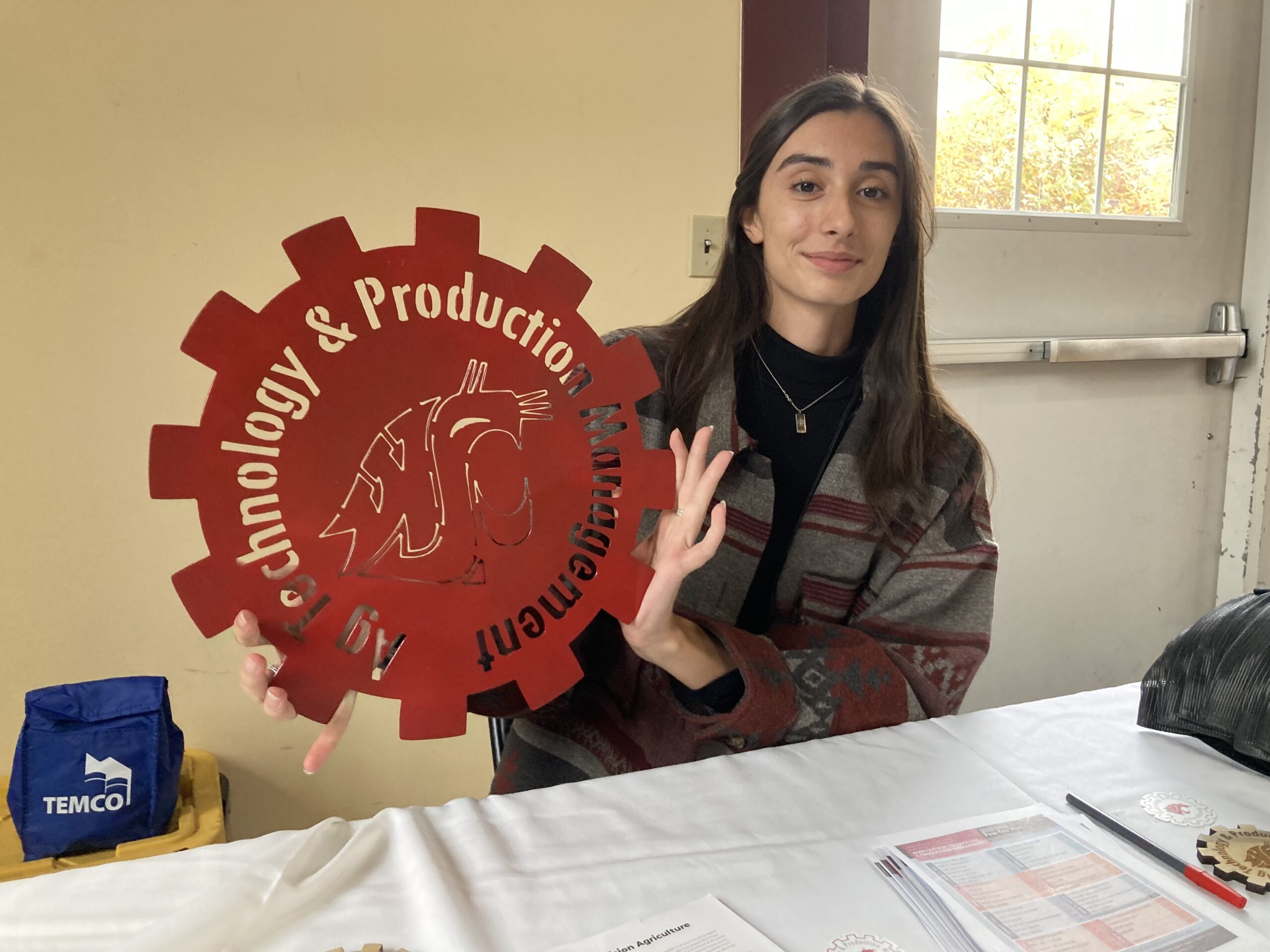 A college student holding up a handmade metal cutout sign in the shape of a gear that says "Ag Technology & Production Management"