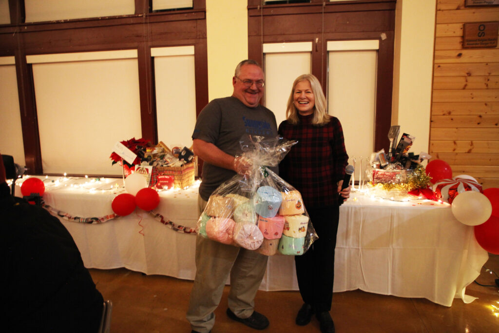 The winner of the toilet paper rolls gift basket beams brightly as Nancy Deringer hands it over in front of the raffle table.