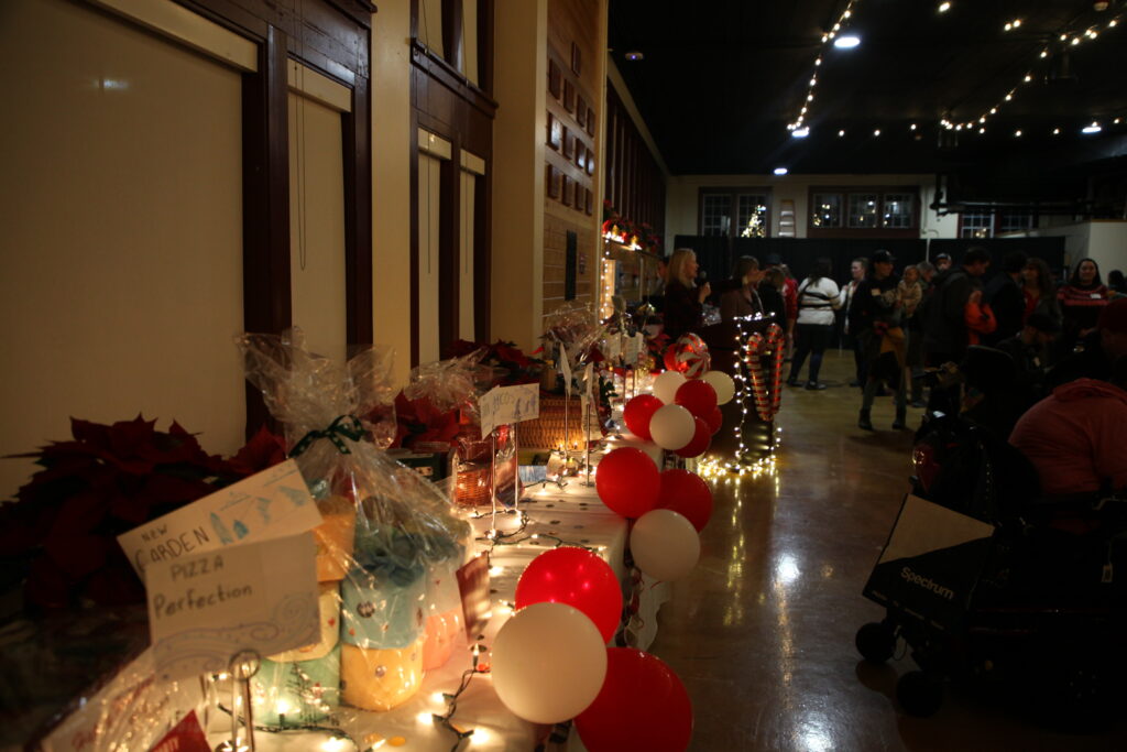 A table of gift raffles is decorated with confetti, christmas lights, poinsettias, and balloons. Some of the raffle items are a Pizza Perfection and a New Garden raffle, and one is a giant package of about 25 toilet paper rolls wrapped beautifully.