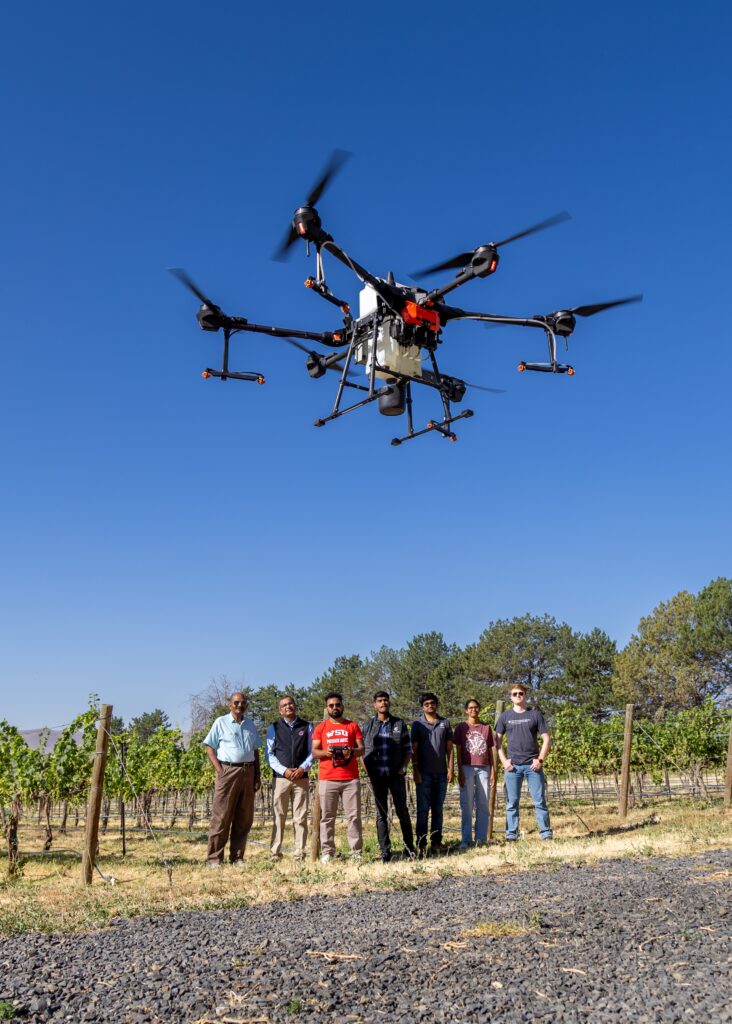 In an orchard setting, a drone flies above a group of observers, one holding a remote control.