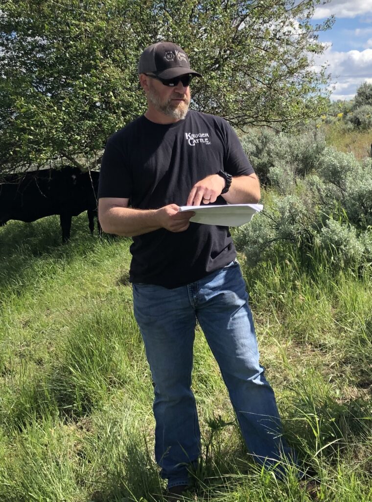 Chad Kruger holds important papers, sporting his Kruger Cattle LLC shirt and matching hat. He stands in the grass, a cow and tree and bushes behind him.