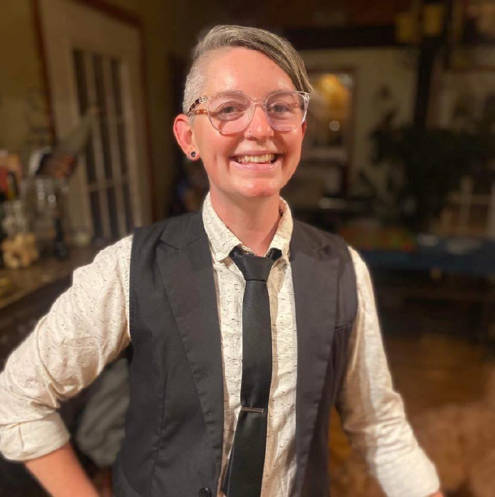 Rae Olsson poses in their home, wearing a professional suit-inspired outfit, a big smile on their face.