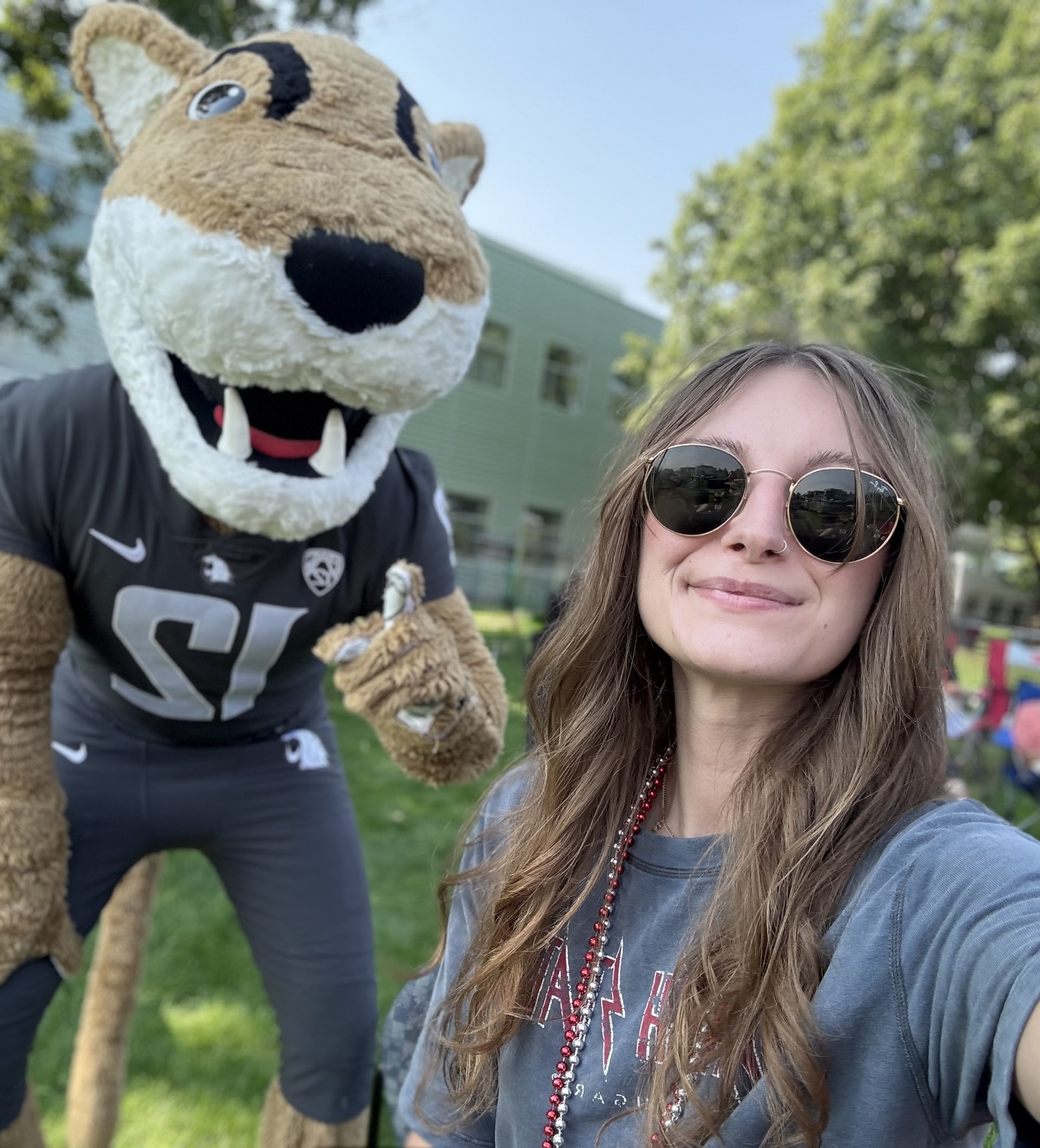 Mackenzie Noyola, wearing sunglasses, WSU-colored beads, and a crimson and gray tshirt, poses with Butch.