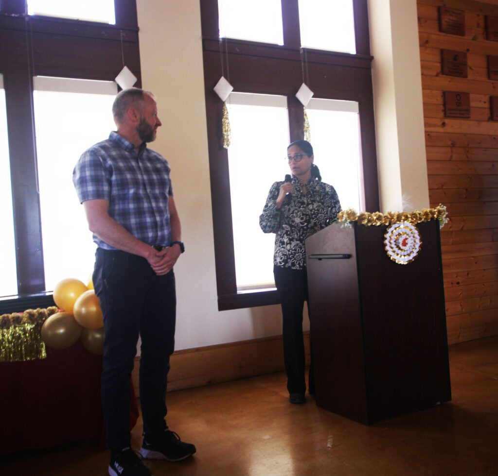 An awardee stands by the certificates table at Faculty and Staff Awards, their nominator speaking about them at a podium.