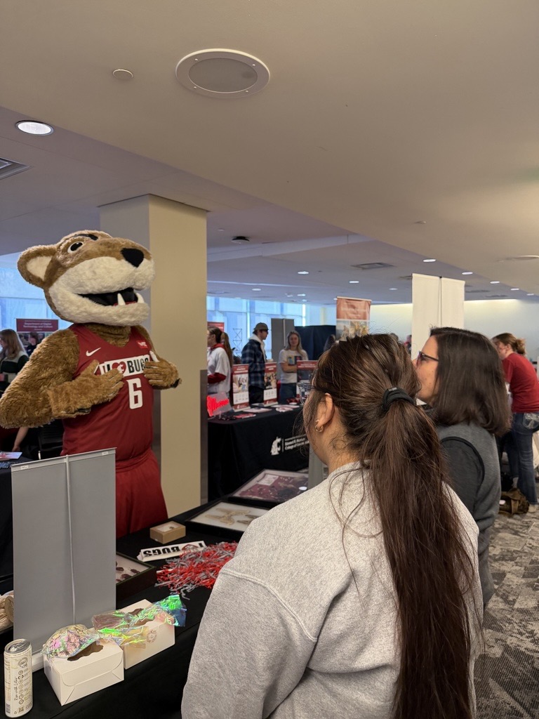 Butch has an I Love Bugs sticker that he is showing off to a student and an advisor at a tabling event.