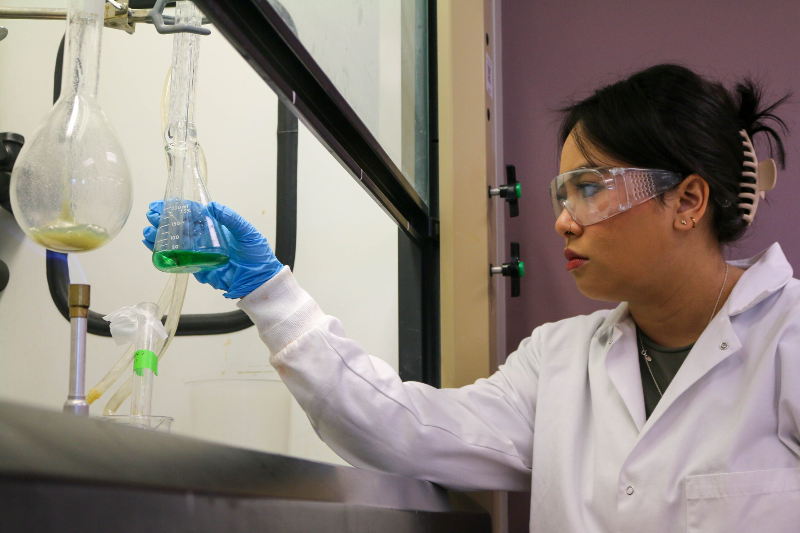 Person wearing a lab coat and goggles holding examining a beaker of content.