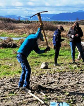 Person holding a mattock about to swing into the ground, some people in the background surrounded by farm field and mountains