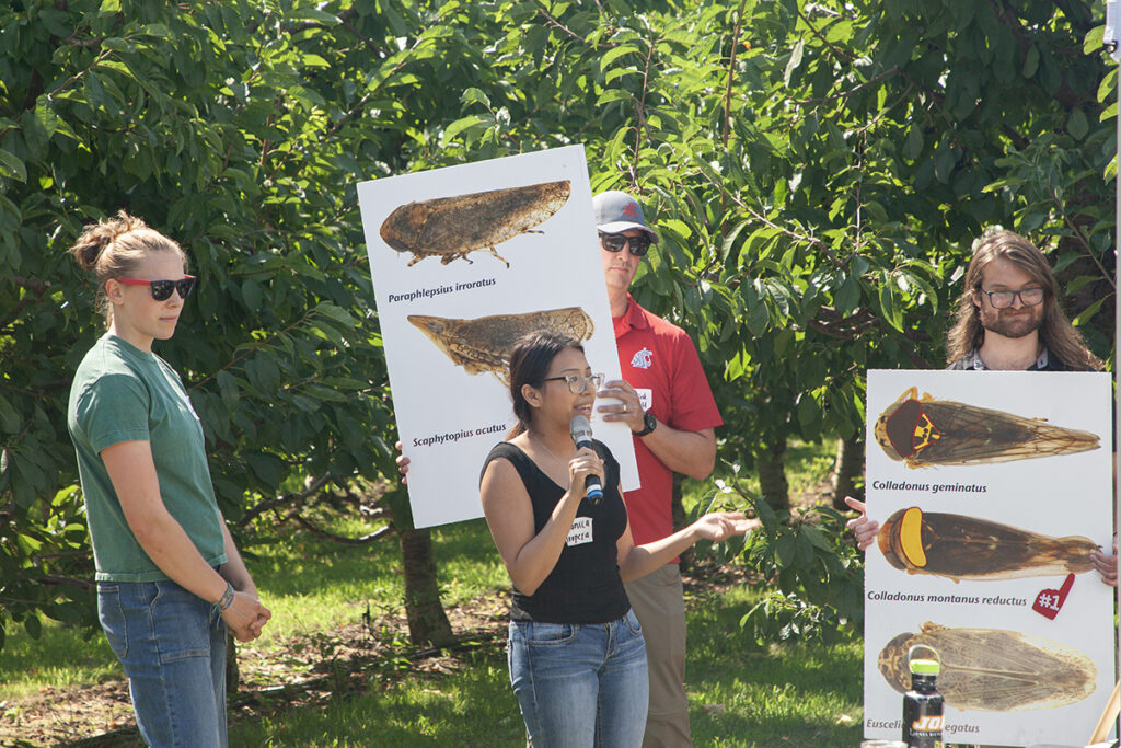 Female undergraduate researcher speaking with microphone while two male researchers hold signs, and second female undergraduate researchers prepares to speak