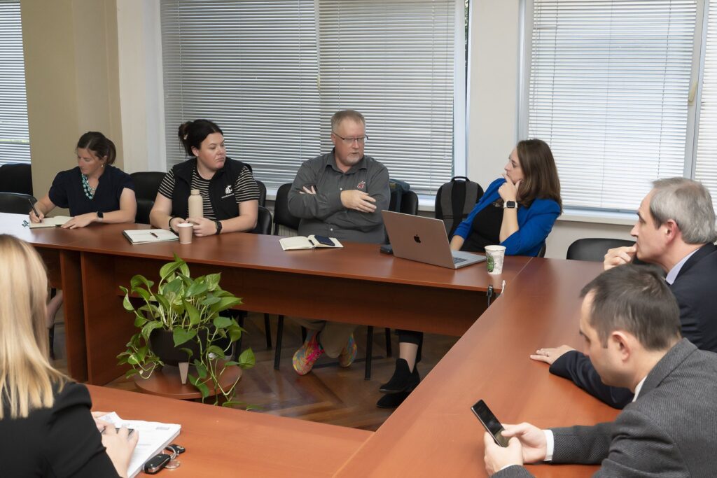 Seven people seated at tables that form a square. One person looks at their phone, one writes on a piece of paper. On has a laptop in front of her. On the table are paper cups, and a water bottle. There is a plant in the area between the tables.