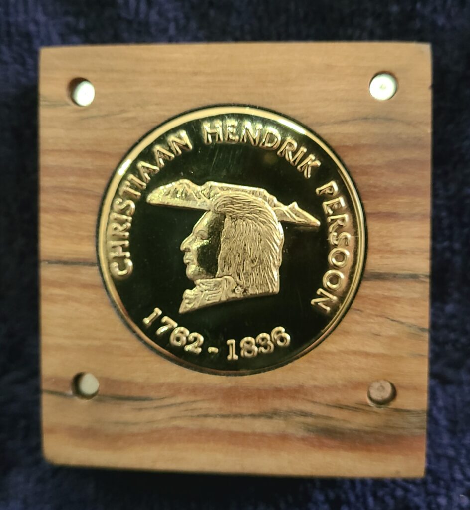 A gold medal embedded in a piece of wood. The medal reads Christiaan Hendrik Persoon, 1762-1836, with an emblem of a man's head on it.