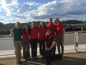 A group of seven people standing against a railing overlooking a river and the city of Pittsburgh in the background.