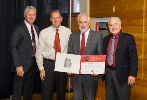 Four men in suits look at camera with Tom Lumpkin holding a mortar board containing his award.