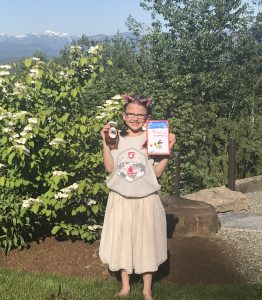 Little girl holds a jar of honey in one and a hand-made book in the other, standing in front of a flowering tree with mountains in the background.