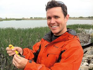 Owen holds a yellow piece of plastic and wears an orange jacket with a pond on the background.